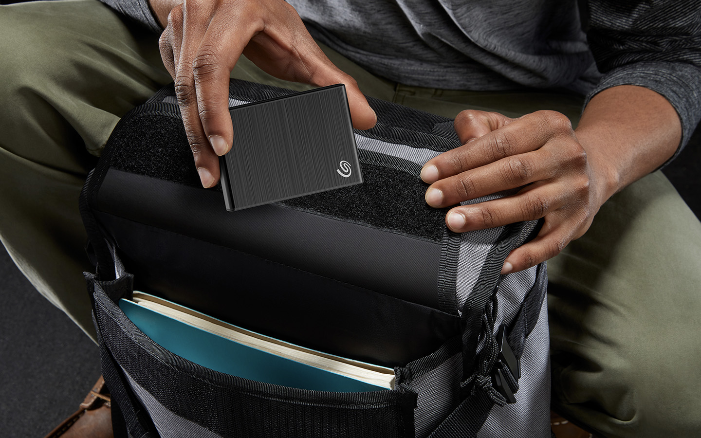 Close-up of two hands above a gray backpack placing a Seagate compact, portable One Touch hard drive in a pocket.