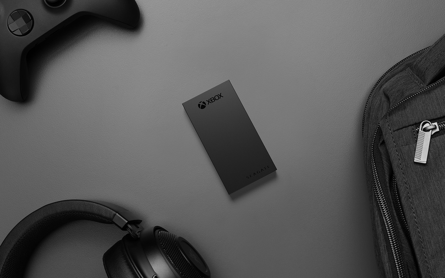 Black Seagate external SSD for Xbox is surrounded by a black Xbox controller, gaming headphones and backpack.