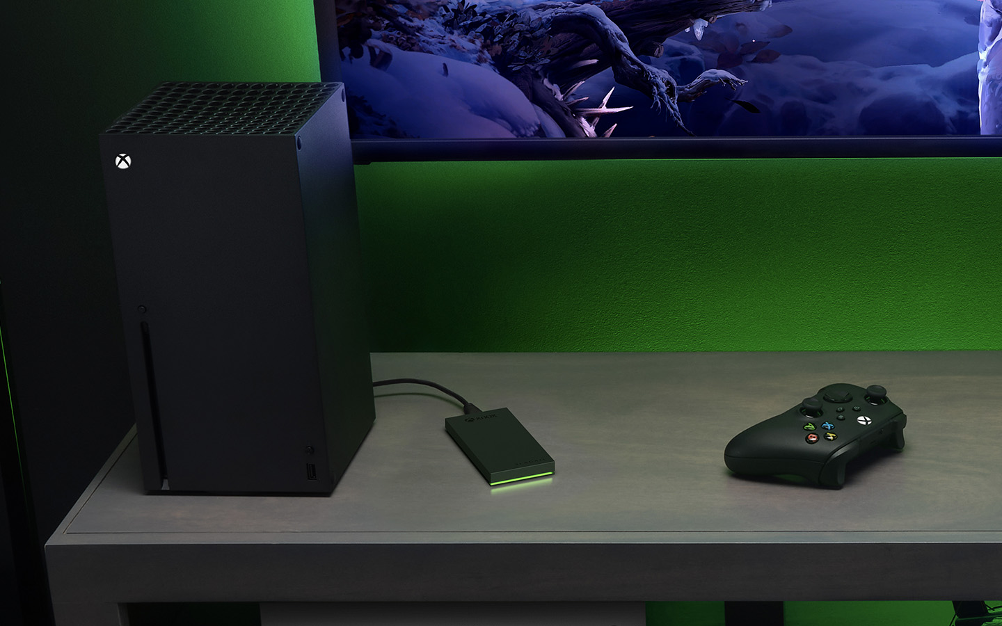 Black Xbox gaming tower with a black Seagate for Xbox external gaming drive stands on a table next to a handheld device and graphics screen.