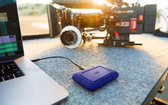 LaCie Rugged SSD Pro5 connected to a laptop sits in front of a RED digital cinema camera, suggesting video production or data transfer.