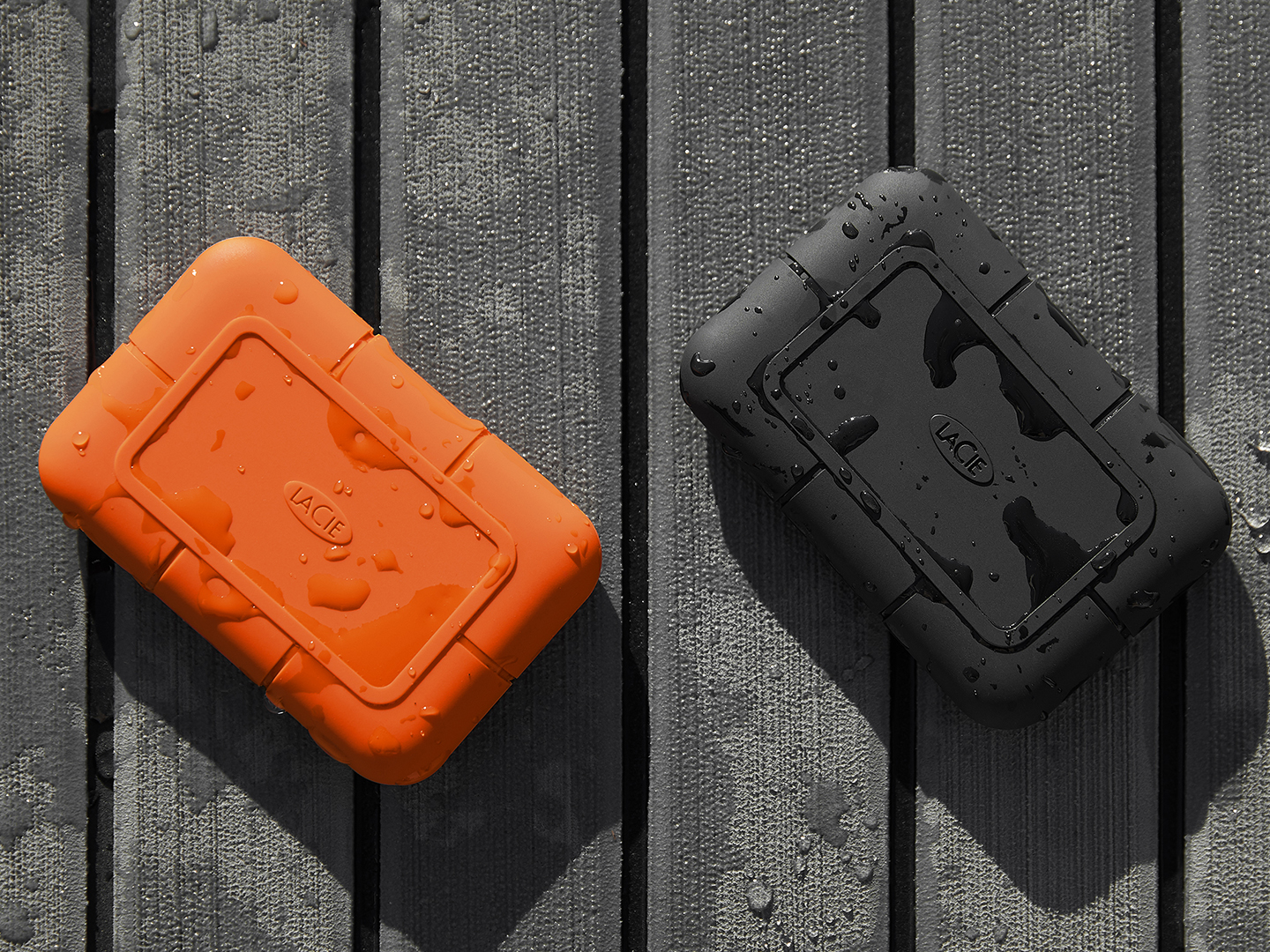 Two wet LaCie Rugged Mini drives — one orange and one black — rest on an outdoor table, highlighting their durability in harsh conditions.