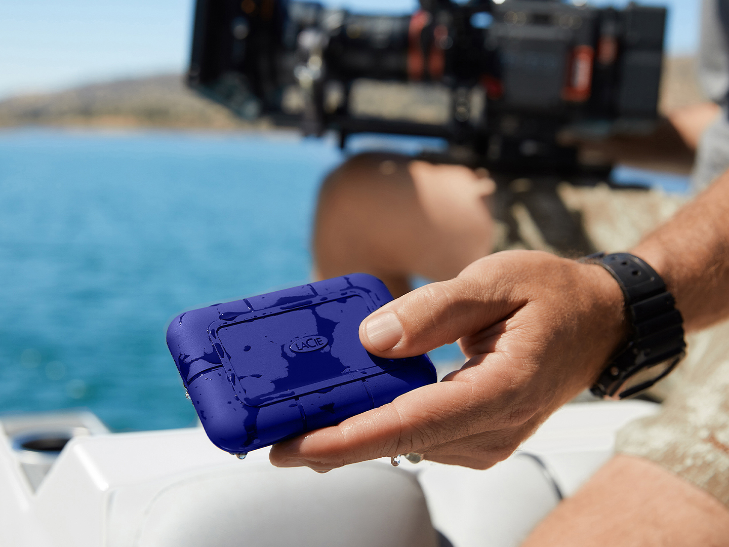 Person in boat holds wet LaCie Rugged SSD Pro5 and large video camera, showing its compact size and rugged durability. 
