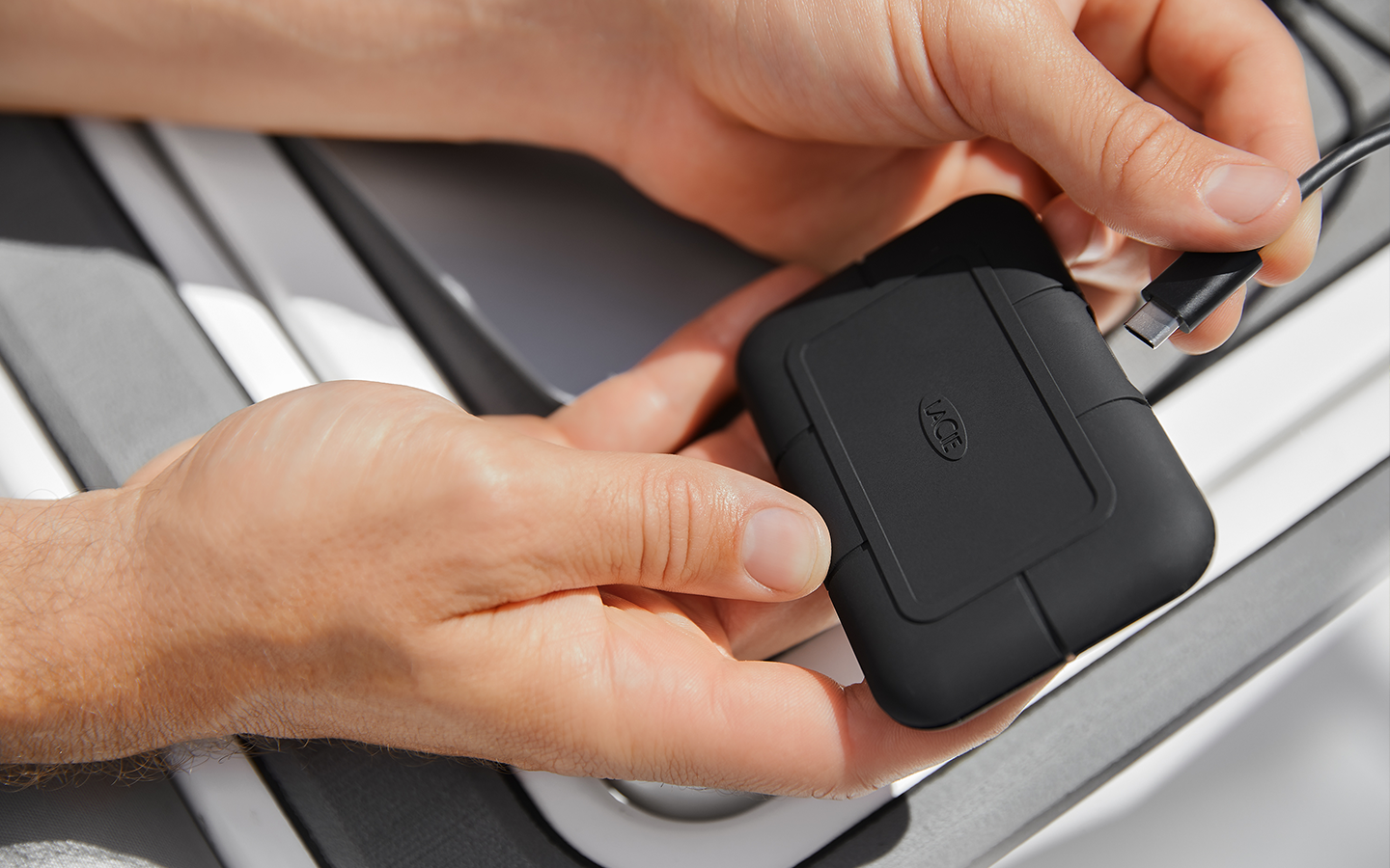 Close-up of hands connecting a LaCie Rugged SSD Pro with a cable; the drive is held in one hand and plugged in with the other.