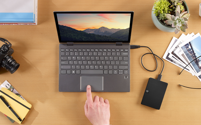 Person using a laptop trackpad is surrounded by a camera, printed photos, and connected Seagate portable hard drive.