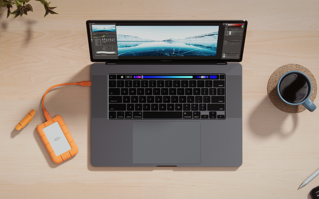 Overhead shot of a laptop computer plugged into a LaCie Rugged Mini SSD sits on a desk next to a cup of coffee.