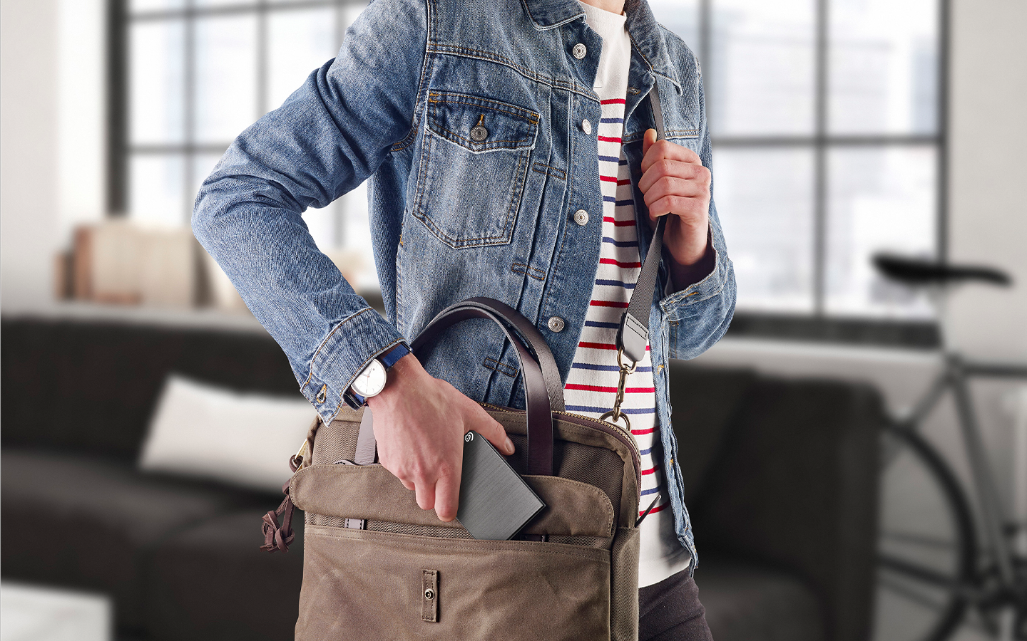 Person puts a Seagate portable external drive into the open pocket of a gray messenger bag being carried.