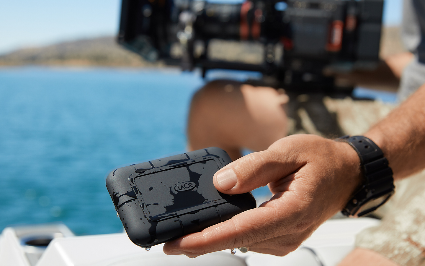 Person next to water holds a wet LaCie Rugged SSD Pro5 in one hand and a large video camera in the other.