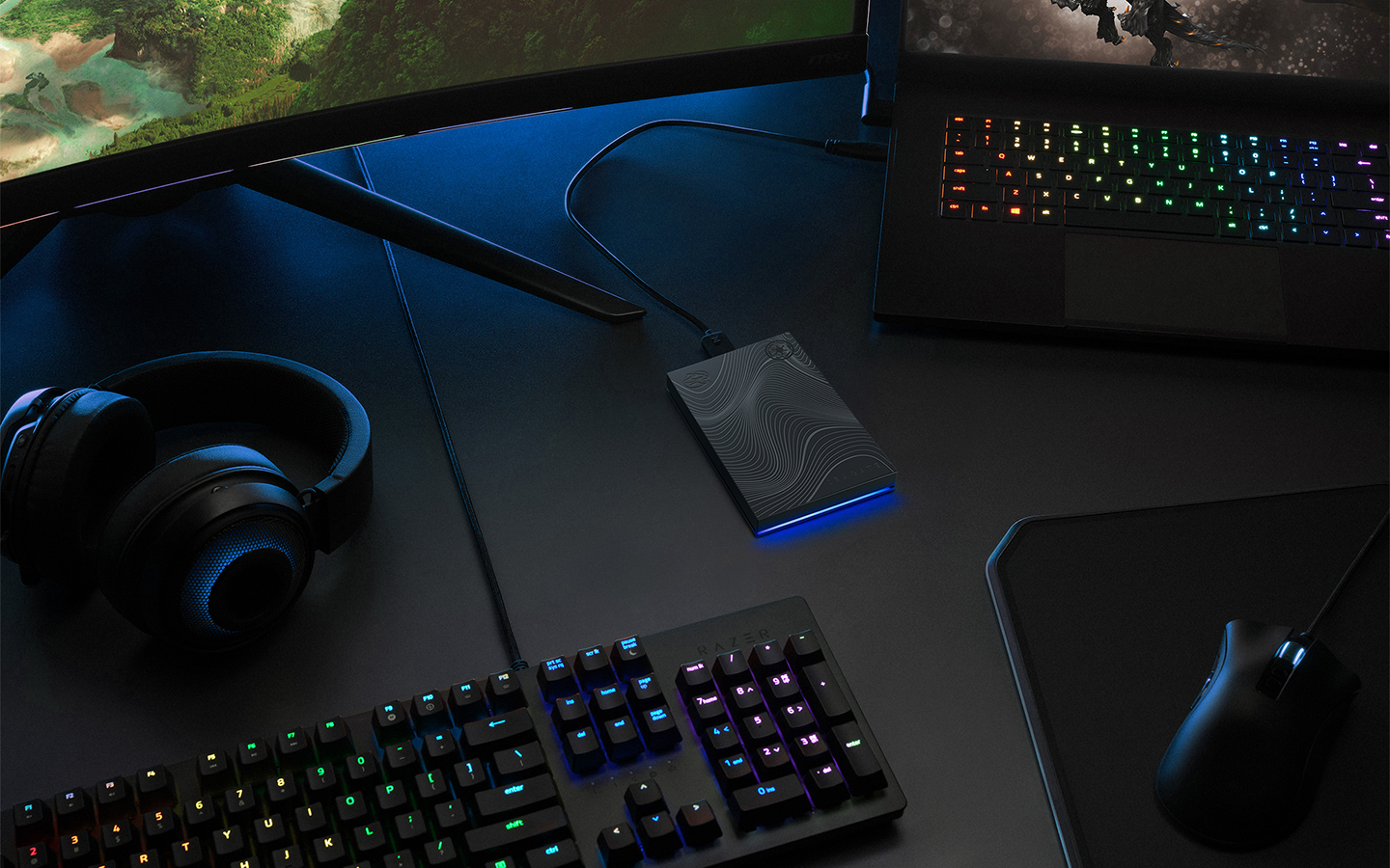 Seagate mini portable drive sits at the center of a gaming laptop, mouse, keyboard, and headphones.