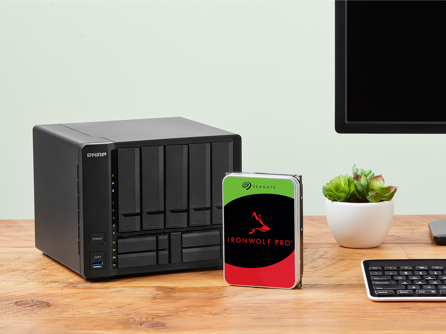 IronWolf Pro drive positioned centrally on a desk is surrounded by a NAS system, computer screen, and keyboard.