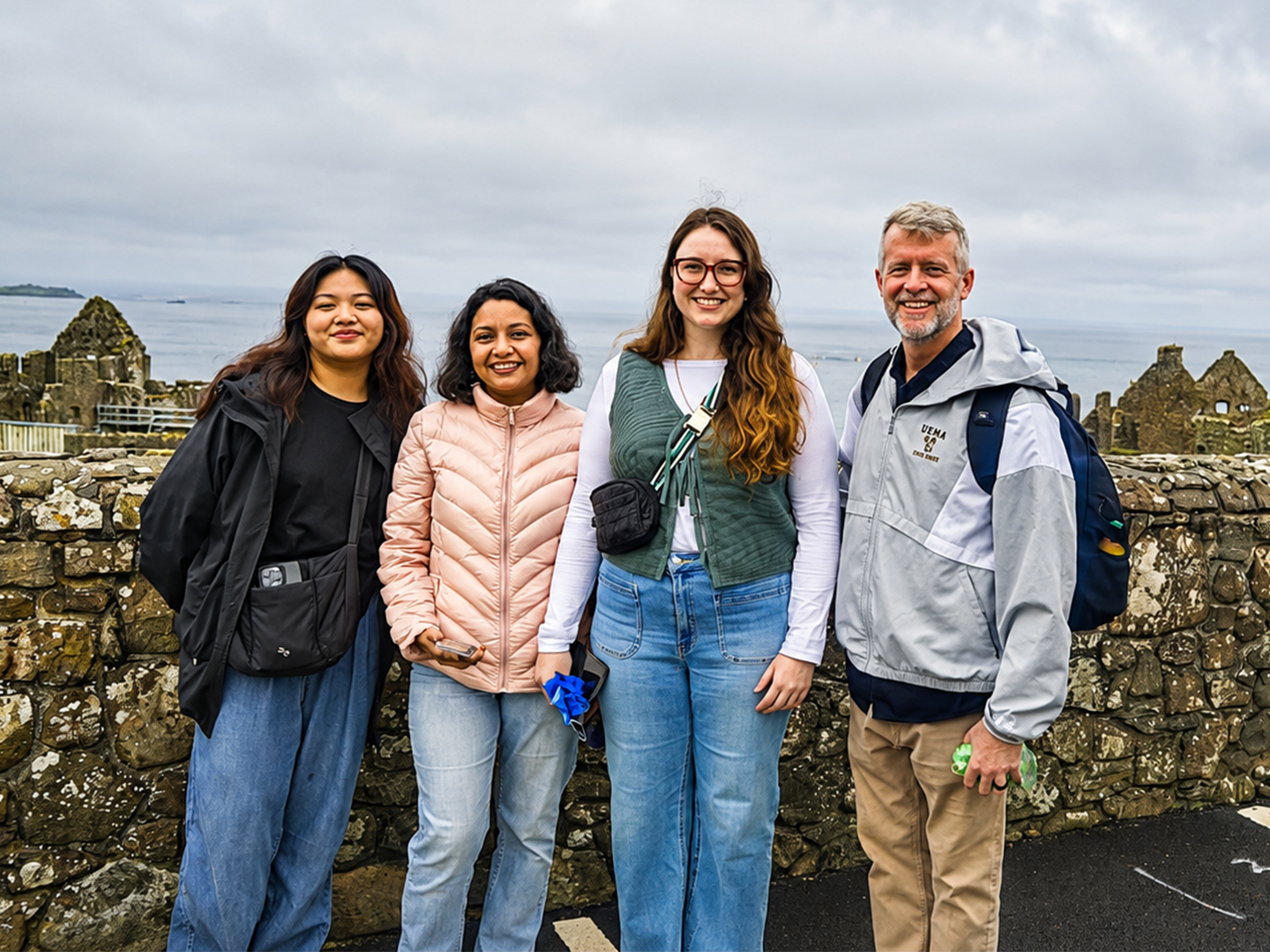 Four people pose outdoors in front of a stone wall with the sea visible in the distance.