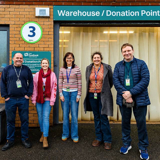 Five people stand outside a building entrance beneath a sign reading “Warehouse/Donation Point.” 