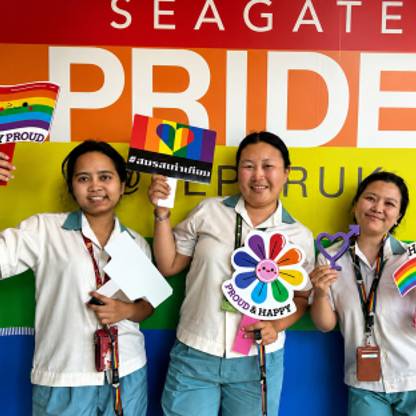 Three people pose in front of a Seagate Pride display while holding Pride-themed signs and decorations. 