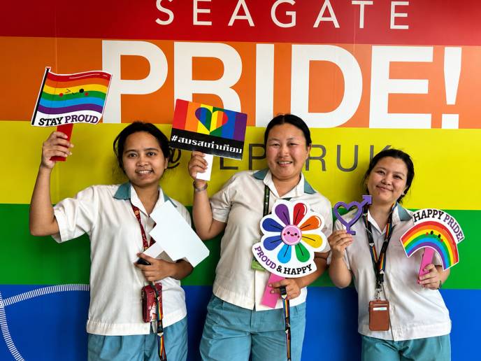 Three people pose in front of a Seagate Pride display while holding Pride-themed signs and decorations. 