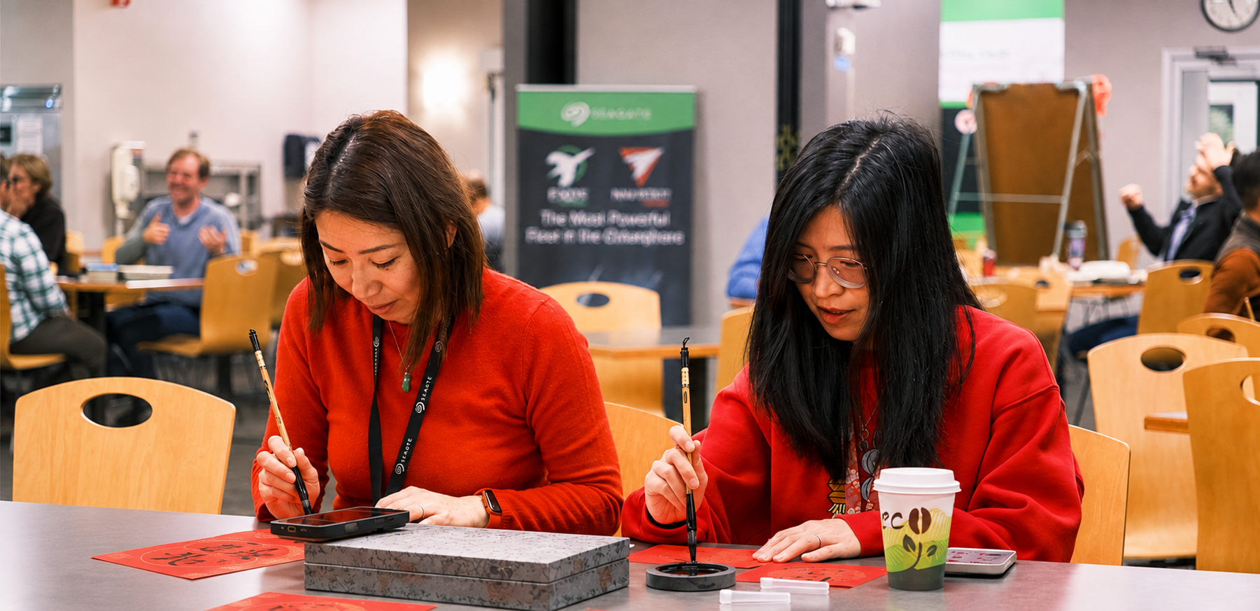 Two people in red shirts sit at a table and use paint brushes to work on a project.