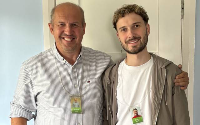 Shaun and his son Harry standing side by side indoors, wearing ID badges, one with an arm around the other, facing the camera.
