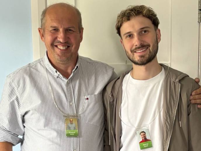 Shaun and his son Harry standing side by side indoors, wearing ID badges, one with an arm around the other, facing the camera. 