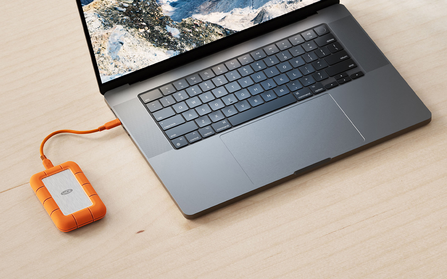 Laptop on wood surface showing aerial landscape on screen; LaCie Rugged SSD4 with orange casing connected.
