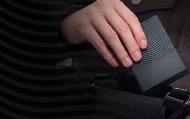 Close-up of a hand holding a black Seagate portable drive over a bag.