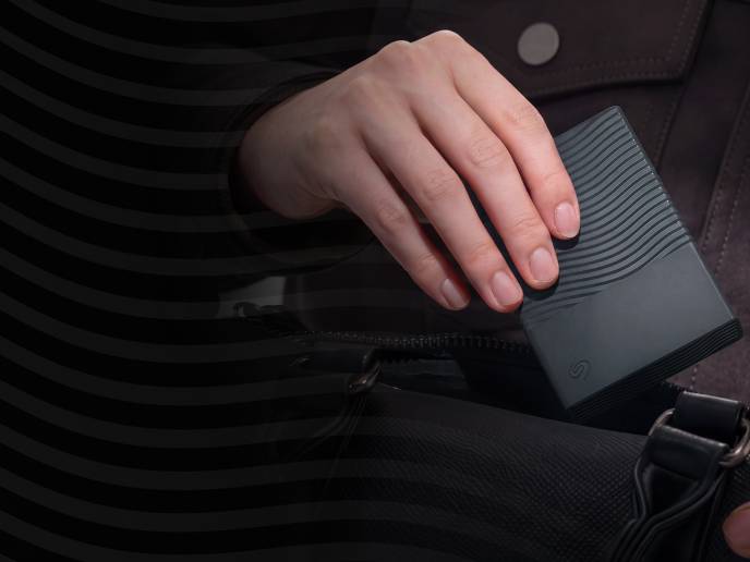 Close-up of a hand holding a black Seagate portable drive over a bag. 